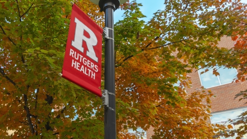 A Rutgers Health banner hangs on a light pole on campus with trees in the background