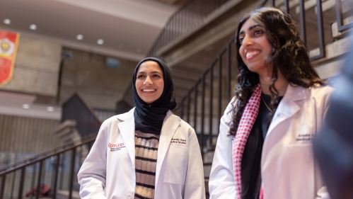Two medical students in white coats smile and talk with others in the Great Hall
