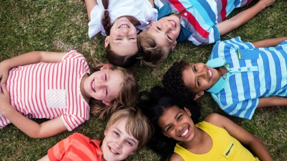 An aerial image of 6 smiling children laying on the grass with their heads in a circle.