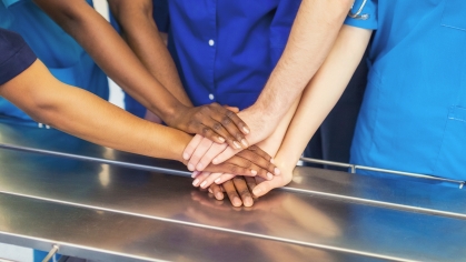 Doctors wearing blue scrubs stack their hands to signify teamwork