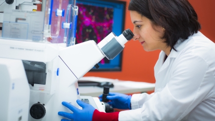 A researcher wearing a white lab coat and blue exam gloves peers into a microscope