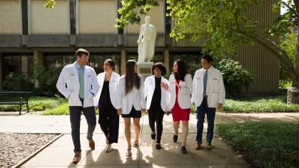 A diverse group of medical students wearing white coats walk on campus in front of a statue of Hippocrates