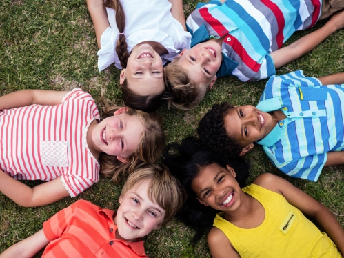 An aerial image of 6 smiling children laying on the grass with their heads in a circle.