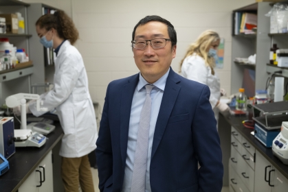 Dr. Hu stands in a lab; two researchers work at benches in the background