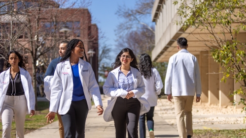 Students in white coats walk outside on campus near the Hippocrates statue